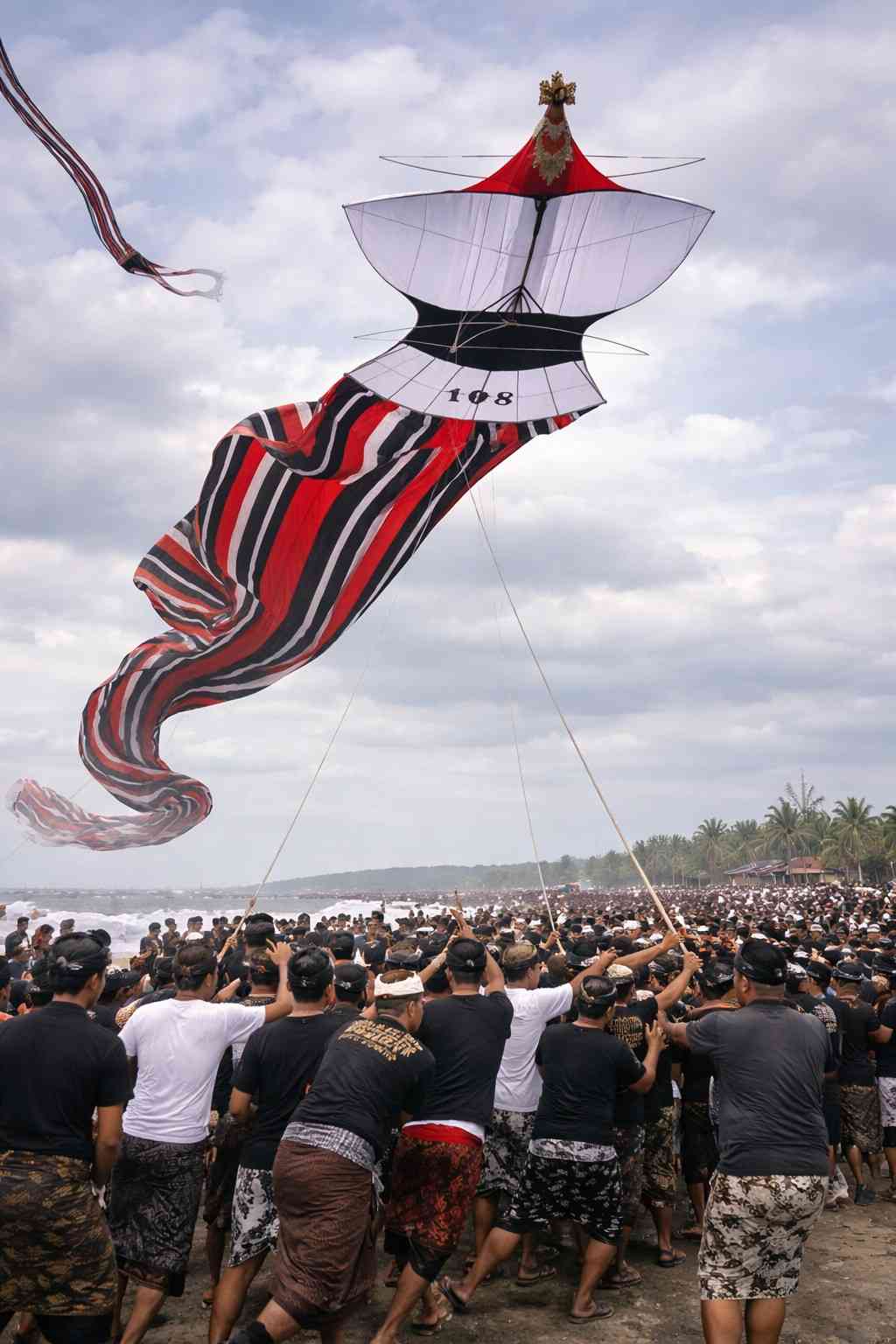 ayangan janggan raksasa khas Bali diterbangkan oleh warga dengan pakaian adat di Pantai Padang Galak saat Festival Layang-Layang Bali, dengan ekor panjang bermotif merah hitam dan langit cerah di siang hari.